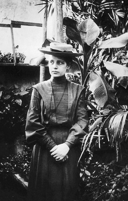 Lise Meitner around 1906 in Vienna. She is wearing a long dress and a hat, standing among large plants and looking to the side. Copyright: public domain  