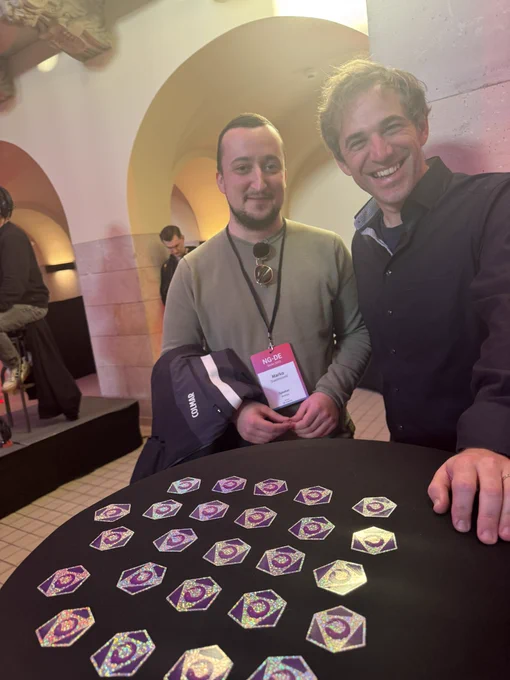 Two smiling people stand by a round table covered with shiny hexagonal stickers featuring a purple design. They appear to be at a conference, with one wearing a badge labeled “NG-DE.” The setting has warm lighting, arched walls, and a casual atmosphere.