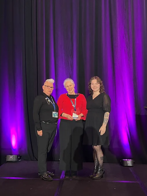 A photo of Jenny Hope (Left), Carolyn McDougall, award winner holding her award (centre) and Lila Jorgenson (Right) in front of a purple and black background