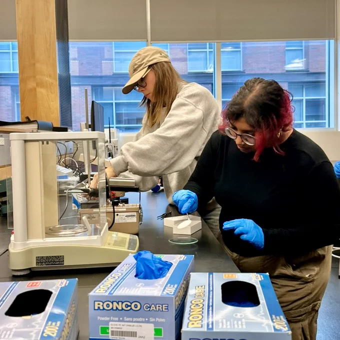 Two students in a lab at Ontario Tech’s north campus. They are standing next to a table working with lab equipment. The student toward the front of the frame is wearing blue latex gloves. 
