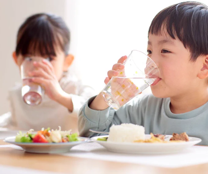 Two children sitting at a table, each drinking from a clear glass of water. 
