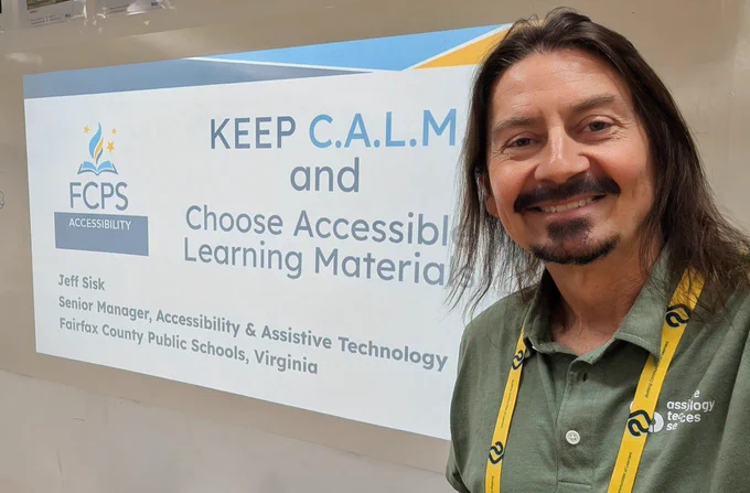 Jeff is standing next to an electronic presentation board that reads "Keep CALM and Choose Accessibile Learning Materials".