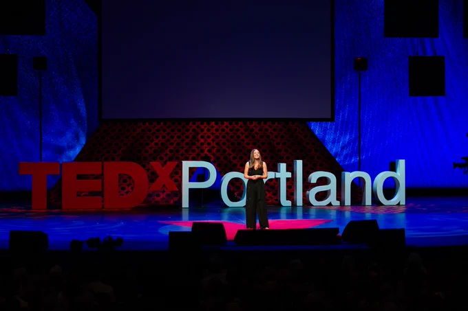 A person stands on stage speaking at a TEDxPortland event, with the event's large red logo displayed prominently in the background.