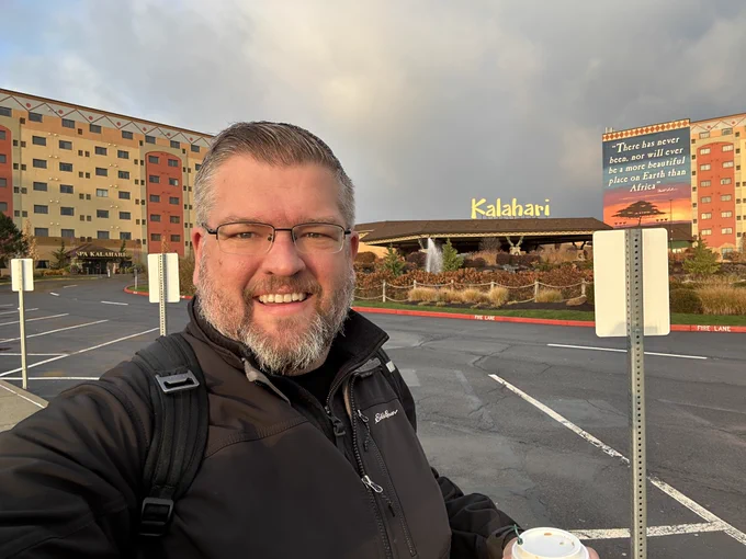 Smiling man with glasses and backpack holding coffee in a hotel parking lot, Kalahari resort sign and multi-story building behind him at sunrise.