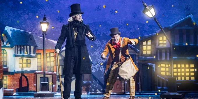 Two characters from "Christmas Carol Goes Wrong" performing on stage with a festive, snowy village backdrop. One character appears as Ebenezer Scrooge in a formal outfit and top hat, and the other as the jovial Mr. Fezziwig in a colourful costume and bowler hat.