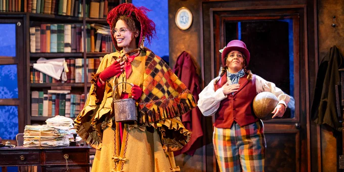 Two actors on stage in a theatrical setting, with one dressed in a vibrant patchwork coat and wild red hair, and the other in a brown hat and plaid trousers, both surrounded by books and props, conveying a whimsical scene.