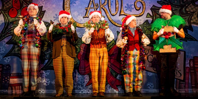 Five performers in festive attire on stage displaying Christmas-themed costumes, including Santa hats, wreaths, and a tree outfit, with a backdrop labeled 'CAROL'.
