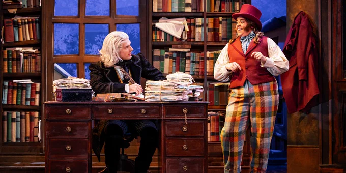 A scene from "Christmas Carol Goes Wrong" featuring a character resembling Ebenezer Scrooge seated at a desk piled high with papers, while another character, likely playing Bob Cratchit, stands beside the desk dressed in colourful attire, both talking in a room decorated to look like a 19th-century office.