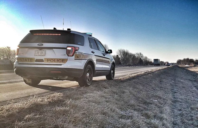 A Ford State Police SUV parked on the side of a highway during sunset, with the road and distant traffic visible in the background.