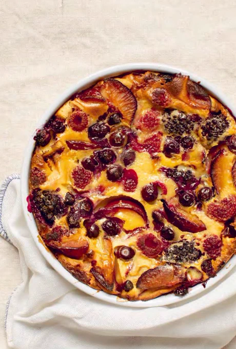 A golden-brown French clafoutis in a round 10-inch baking dish, topped with a mix of red fruits such as cherries, raspberries, and strawberries, with a slightly puffed custard texture, ready to serve.