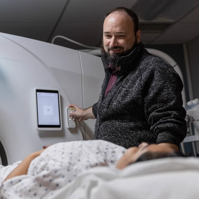 A person operates a medical imaging device while another person lies on an examination table, both in a hospital setting.