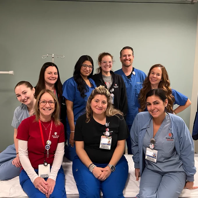 Group of nine hospital staff in scrubs, smiling, posed in a medical room. Some are seated on a hospital bed, others are standing. They all wear identification badges.