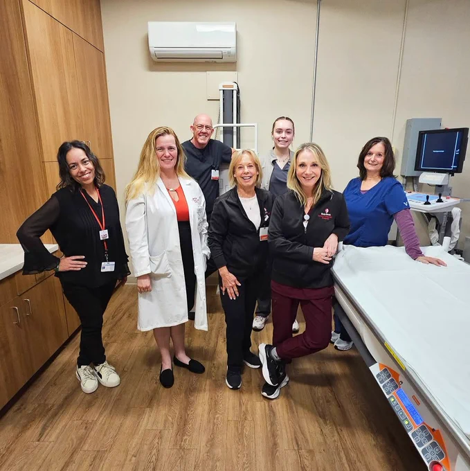 Group of medical staff smiling in a hospital room, including doctors and nurses, some wearing lab coats, scrubs, and badges. The room features medical equipment and a patient bed.