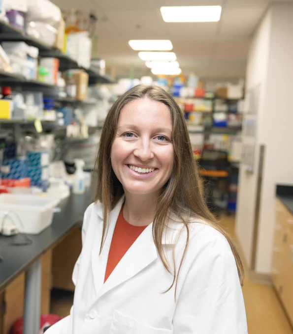 Cali Love sitting at her lab bench, wearing a white lab coat and an orange shirt.