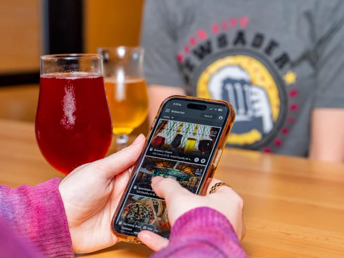 A person at a table browsing a food and drink app on a smartphone, with a glass of red beverage nearby, sitting across from another person in the background.