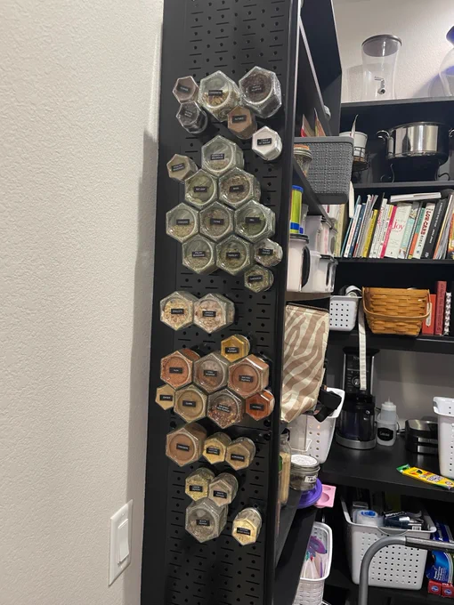 A black Wall Control metal pegboard mounted on a wall, displaying neatly arranged hexagonal spice jars labeled and organized in a vertical kitchen setup.