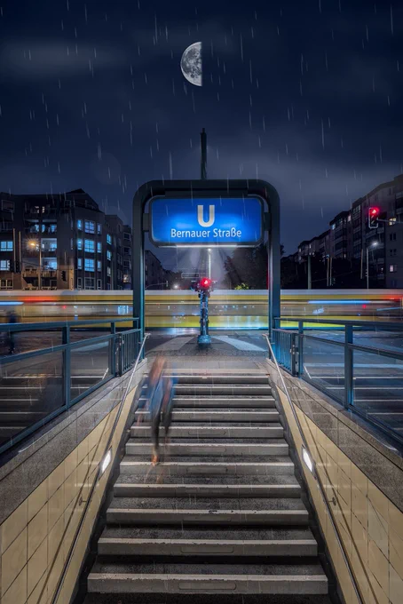 Der Eingang zum Berliner U-Bahnhof „Bernauer Straße“ bei Nacht. Der Blick führt von unten über eine breite Treppe mit Handläufen und beleuchteten Wänden nach oben zur Straße. Oben befindet sich ein großes, rechteckiges Schild mit blauem Hintergrund und weißer Aufschrift: „U Bernauer Straße“. Im Hintergrund ist ein Stadtszenario mit mehrstöckigen Wohnhäusern zu sehen. Eine gelbe Straßenbahn fährt waagrecht durchs Bild, erzeugt durch Langzeitbelichtung eine Bewegungsunschärfe. Eine Person ist auf der Treppe unscharf zu erkennen, vermutlich in Bewegung nach unten. Es regnet leicht, Regentropfen sind sichtbar. Am Himmel steht ein halb beleuchteter Mond, teilweise von Wolken verdeckt. Die Szenerie ist durch Straßenlaternen und Lichtreflexionen auf dem nassen Boden illuminiert.