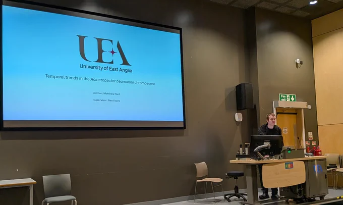 Speaker Matthew Neil at the podium presenting his talk titled "Temporal trends in the Acinetobacter baumannii chromosome". The UEA logo is depiceted above and below is the text "Author: Matthew Neil, Supervisor: Ben Evans".