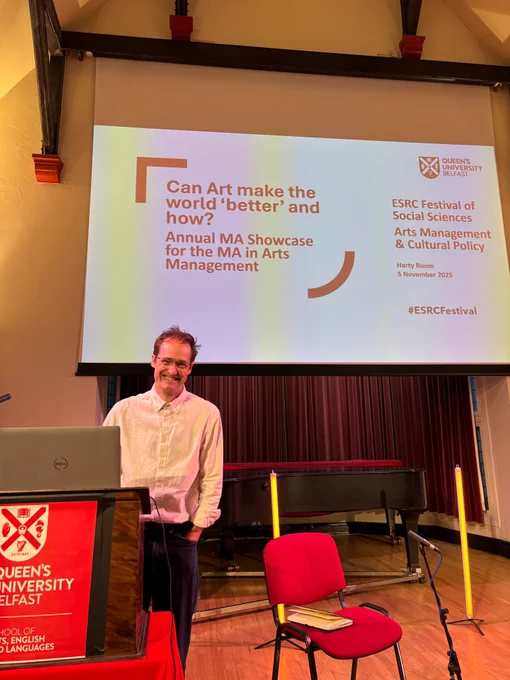 Anthony Schrag wears a white shirt and stands behind a lectern on a stage. Behind him is a screen showing a powerpoint with the talk title 'Can Arts make the world 'better' and how?'