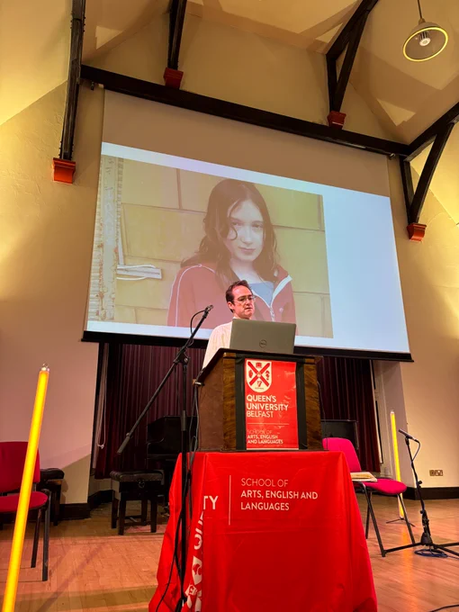 Anthony Schrag speaks at an event in the Harty Room. He is pictured wearing a white shirt standing at a high lectern with a red poster saying 'QUB School of Arts English and Languages. Behind him is a screen showing an image from his talk.