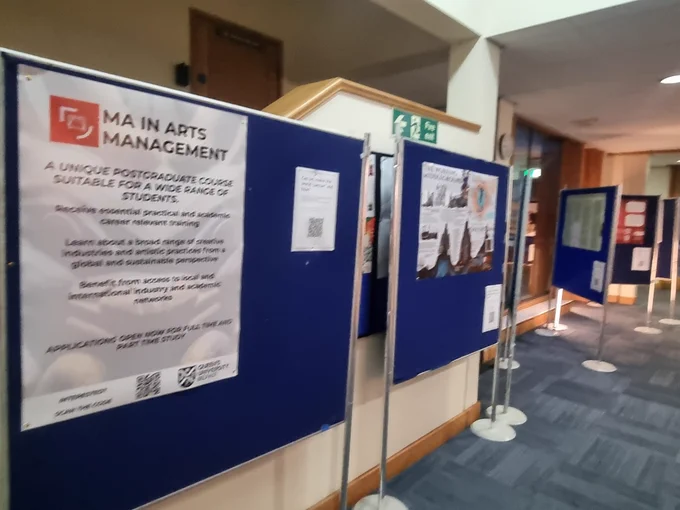 The image shows a series of academic posters from the MA in Arts Management. They are displayed on blue poster stands down a foyer. This event celebrated student's work from the year 2024 - 2025
