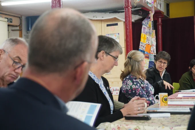 Group of adults engaged in a discussion around a table covered with a floral tablecloth in a cosy room with colourful decorations. Tess White MSP is speaking to a member of the community.