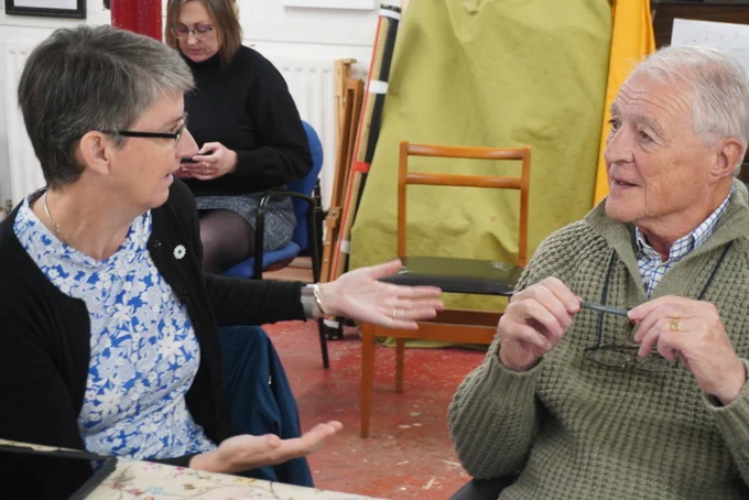 Group of adults engaged in a discussion around a table covered with a floral tablecloth in a cosy room with colourful decorations. Maggie Chapman MSP is speaking to a member of the community.