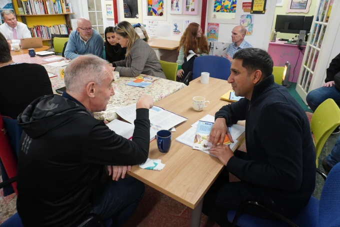 Group of adults engaged in a discussion around a table covered with a floral tablecloth in a cosy room with colourful decorations. Paul McLennan MSP is speaking to a member of the community.