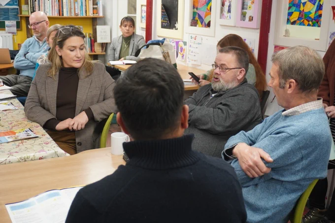Group of adults engaged in a discussion around a table covered with a floral tablecloth in a cozy room with colorful decorations. Karen Adam MSP listens to a discussion.