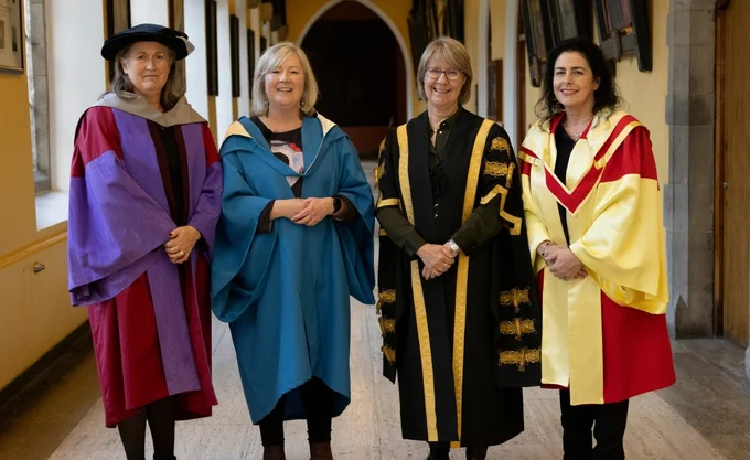 Four women standing side by side in an academic hallway, wearing formal academic robes. The first woman on the left wears a purple and red gown with a black hat. The second woman is in a blue robe, smiling. The third woman wears a black gown with gold embroidery. The fourth woman is in a red and yellow robe, smiling. The hallway features arched doorways and framed pictures on the walls.