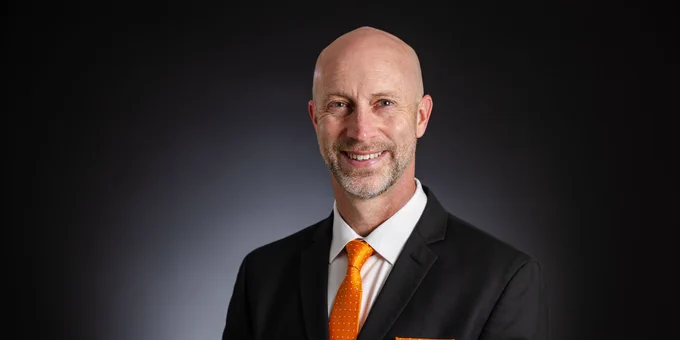 Portrait of a smiling person in a dark suit with an orange tie and pocket square, set against a dark background.