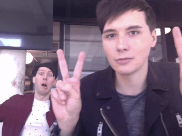 Photo of Dan and Phil sitting at a desk taking a silly selfie gif. Dan is closest to the camera with an emotionless expression, making peace signs with both hands and Phil is sitting further back, making a silly face.
