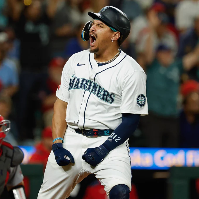 Josh Naylor shouts in celebration. He is wearing a white Seattle Mariners jersey and a dark blue batting helmet.