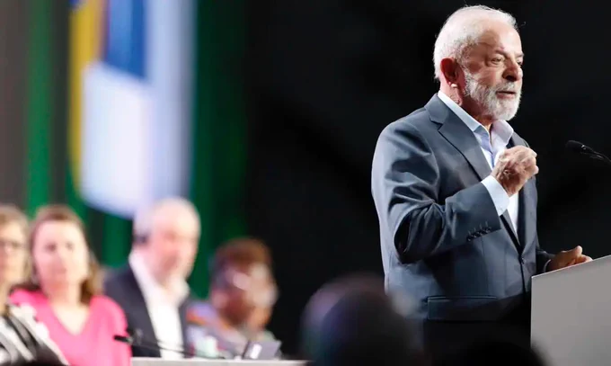 Luiz Inácio Lula da Silva, wearing a grey suit, stands at a podium speaking and gesturing with his right hand. Several other people, including COP30 President André Corrêa do Lago, are seated in the blurred background to his left.