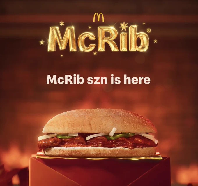 The image is a promotional advertisement for McDonald's featuring the McRib sandwich. Here's a detailed breakdown:


Text Elements:

At the top, the McDonald's logo (golden arches) is visible.
Below it, the word “McRib” is written in a shiny, golden font with sparkles, giving it a festive and celebratory feel.
The tagline reads: “McRib szn is here”, which uses slang ("szn" for "season") to create a casual, trendy vibe.

Visual Elements:

The McRib sandwich is prominently displayed in the center. It consists of:

A long sandwich bun with a slightly textured surface.
Barbecue-sauced pork patty.
Toppings include sliced onions and pickles.


The sandwich is placed on a red pedestal with a golden edge, adding a premium and seasonal touch.
The background is warm and blurred, with tones of orange and red, resembling a cozy fire or festive ambiance.

This image clearly signals the return of the McRib, which is often a limited-time seasonal offering at McDonald's.
