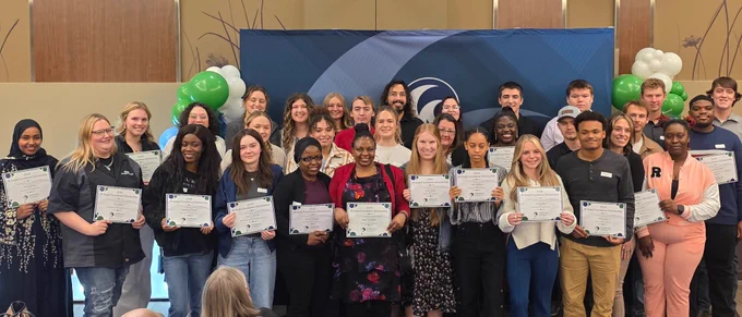 Group of students holding certificates, posing with a teacher in front of a banner with the M State logo, in a room decorated with balloons.
