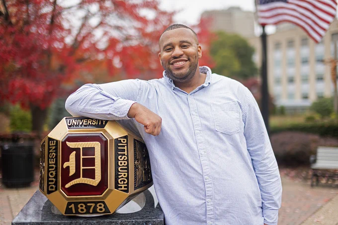 Duquesne alumnus Jeffrey Brown rests his arm over the Duquesne Ring statue on campus in the fall of 2023. He is smiling at the camera. 