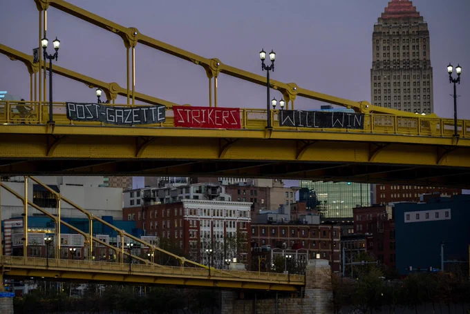 With Downtown Pittsburgh in the background, three banners hang off the Roberto Clemente Bridge, reading "POST-GAZETTE STRIKERS WILL WIN." It is dusk with a soft lavender sky in the background as street lights have begun to turn on along the bridges.