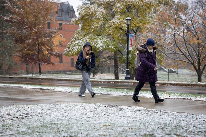 Two Cornellians walk on a snowy path across the Cornell University campus, with one carrying a backpack and the other holding a mobile phone. Historical brick buildings in the background are adorned with autumnal foliage.