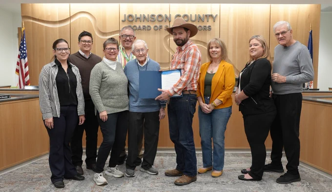 A group of nine people stands together at the front of the county board meeting room for a photo. One person holds a framed proclamation or certificate. They are standing in front of the “Johnson County Board of Supervisors” wall. The group smiles and faces the camera.