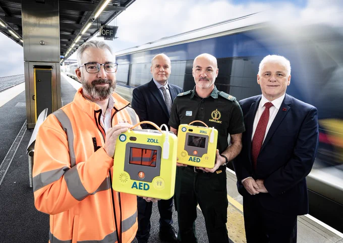 four individuals including a paramedic standing on a train station platform holding two yellow ZOLL AED defibrillators, with a train in the background.