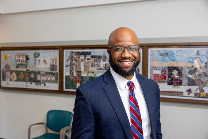 Person with a friendly smile, wearing glasses, a navy suit, and a red-striped tie, standing in an office with colorful mosaic artwork on the wall in the background.