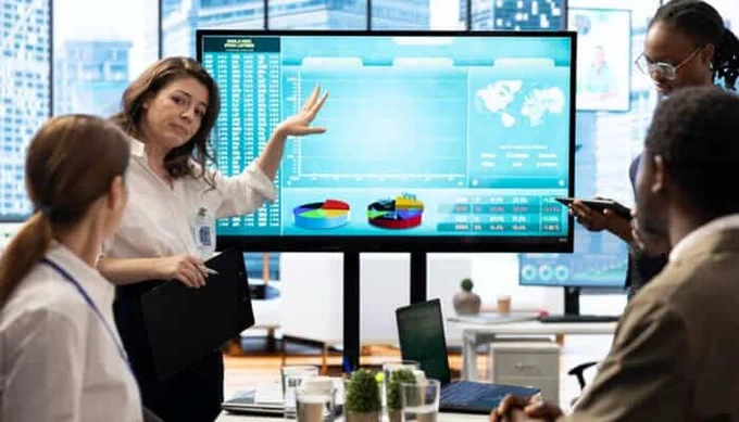 A businesswoman presenting financial charts and data on a large screen to a diverse team during a meeting in a modern office, illustrating data analysis and strategy planning.