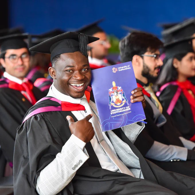 Graduate in gown holding University of Staffordshire programme and smiling at a graduation ceremony.
