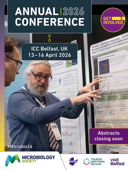 A person gestures while discussing a research poster with another attendee at a conference, with text announcing the Microbiology Society Annual Conference 2026 in Belfast, UK, from 13–16 April.