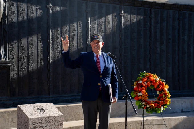 A person in a military cap and dark suit stands next to a wreath at a memorial wall with engraved text.