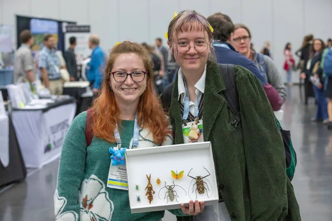 Two people stand indoors at an event holding a display case containing various pinned insects. They are smiling at the camera, and there are several other people and tables in the blurred background.