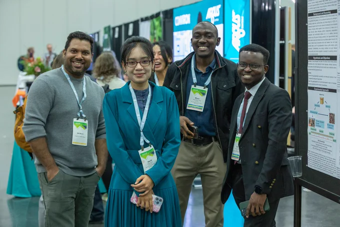 Four people wearing conference badges stand together and smile at the camera in a convention hall, with posters and other attendees visible in the background.