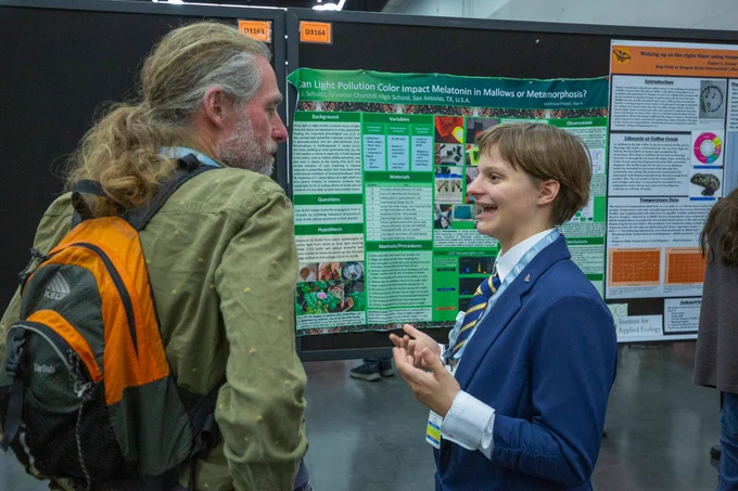 A person in a suit and tie talks to another person with long hair and a backpack in front of a large science poster at an academic conference. Other posters and people are visible in the background.