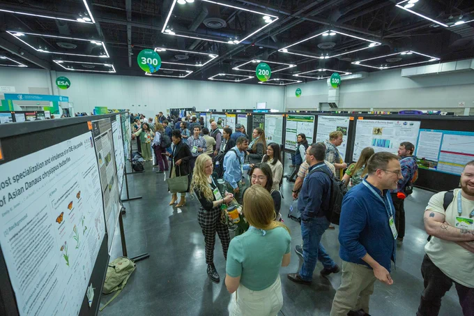 Conference attendees stand and talk in front of research posters displayed on black boards in a large, well-lit hall. Overhead, green signs mark booth numbers, and people are engaged in discussion and presentation.